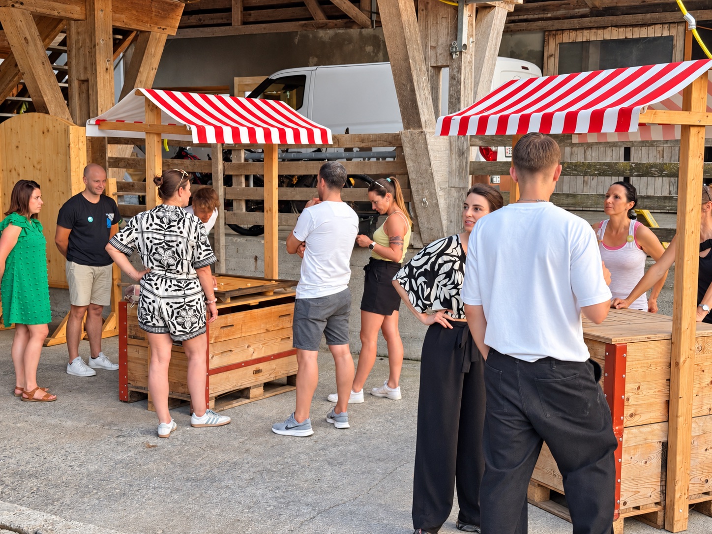 Participants autour de stands de jeux en bois avec auvents rouges et blancs sous une grange